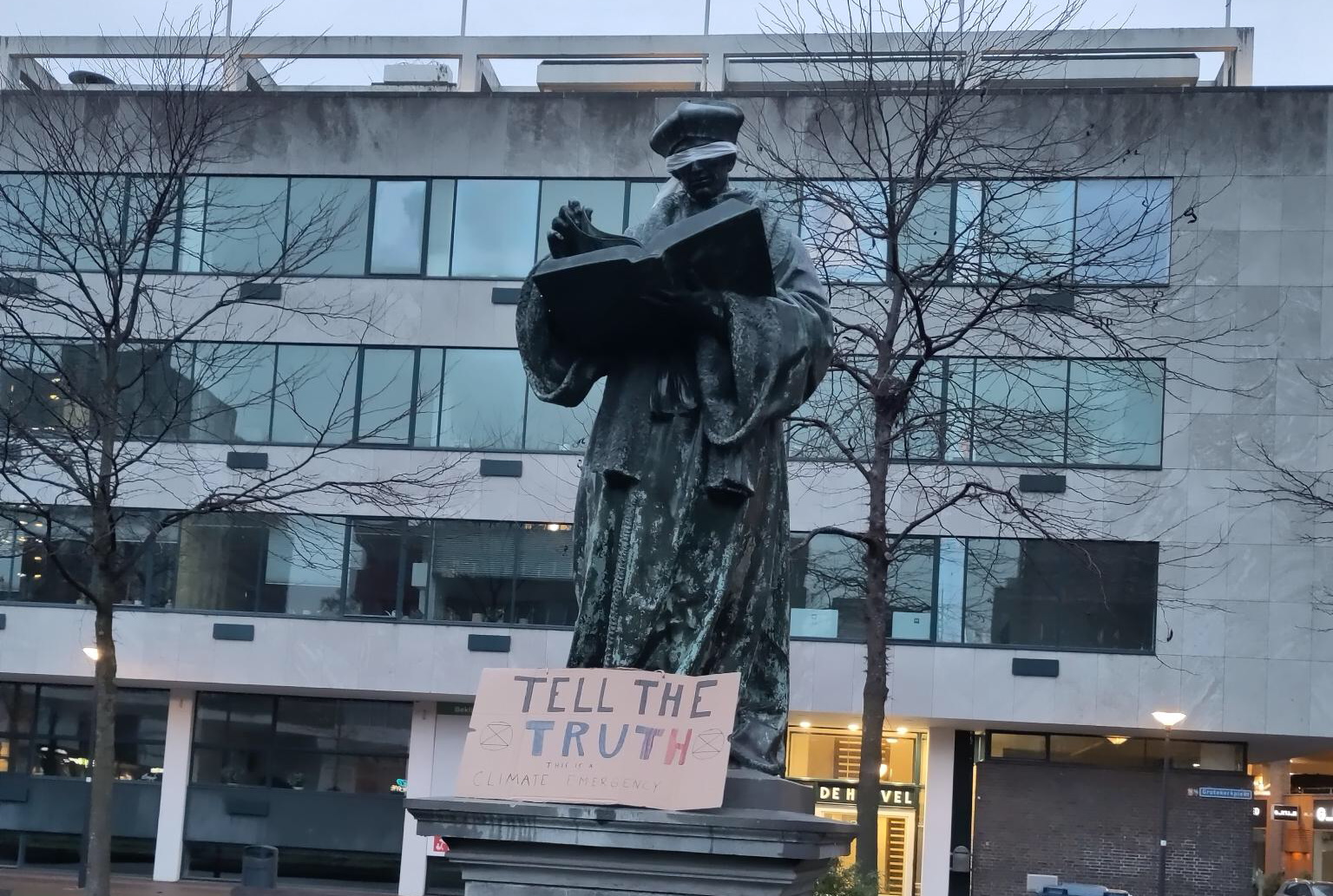 Geblinddoekt Erasmusbeeld op het Grotekerkplein in Rotterdam met daaronder een kartonnen bord met de tekst 'Tell the truth'