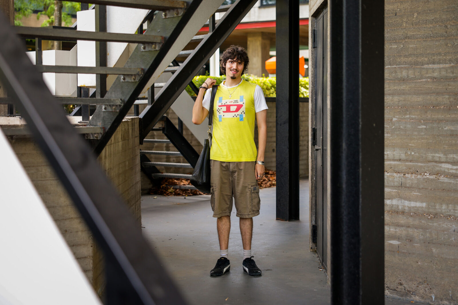 Juan staat onder de trappen aan de buitenkant van het Theilgebouw en draagt een comfortabele korte broek en felgeel shirt met stukjes stof in andere kleuren