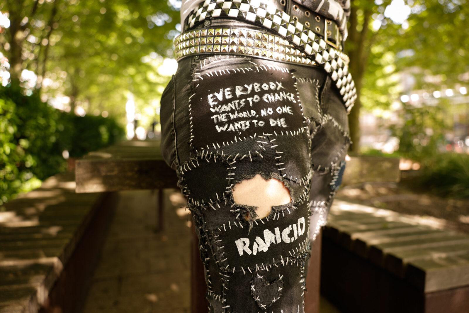 Een closeup van de broek met opgenaaide stukken met daarop teksten als 'Rancid' en 'Everybody want to change the world, no one wants to die'
