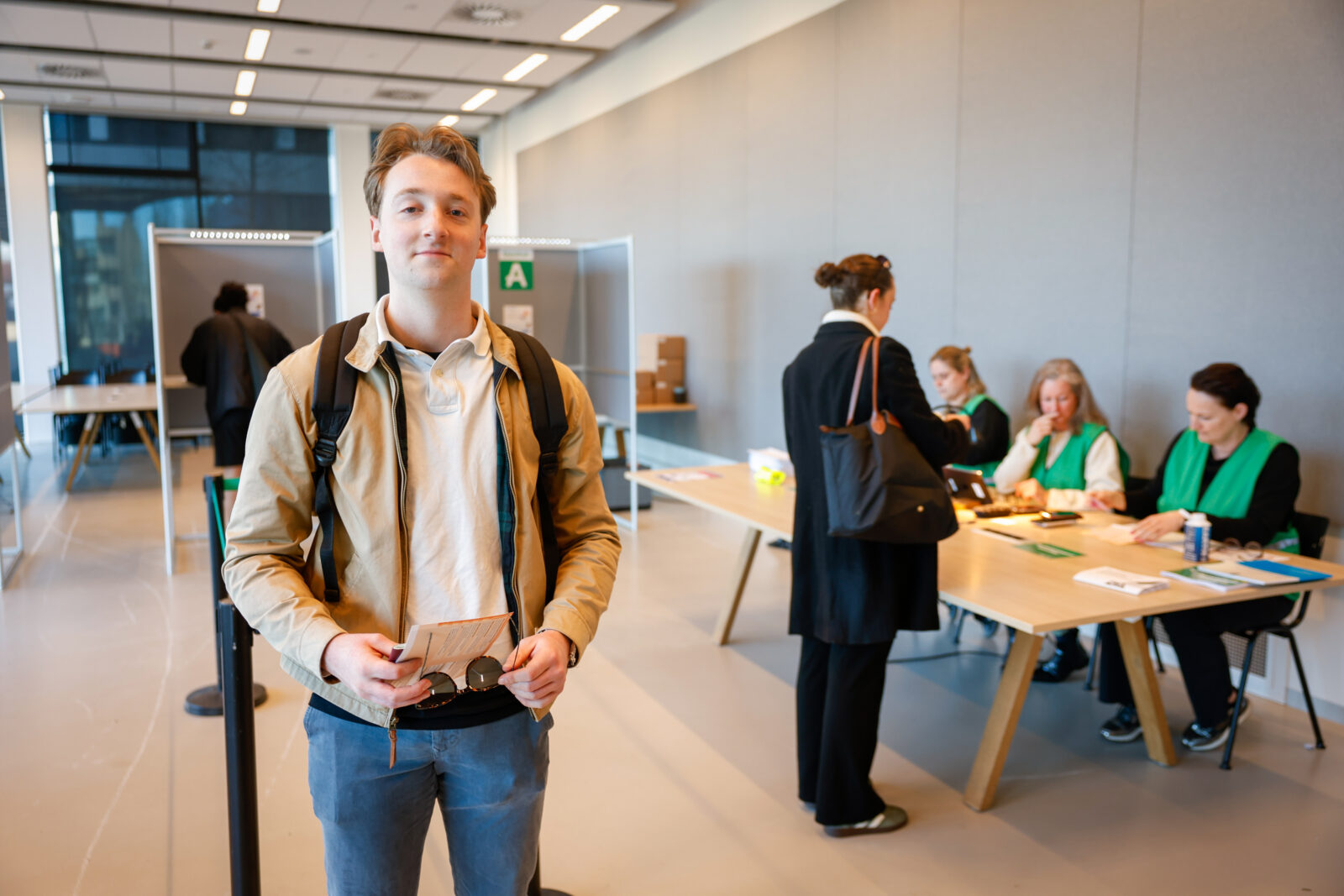 Joris in het stembureau, met op de achtergrond de vrijwilligers en iemand die een stem uitbrengt